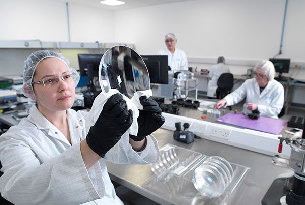 woman wearing white coat in clean room holding up a large lens